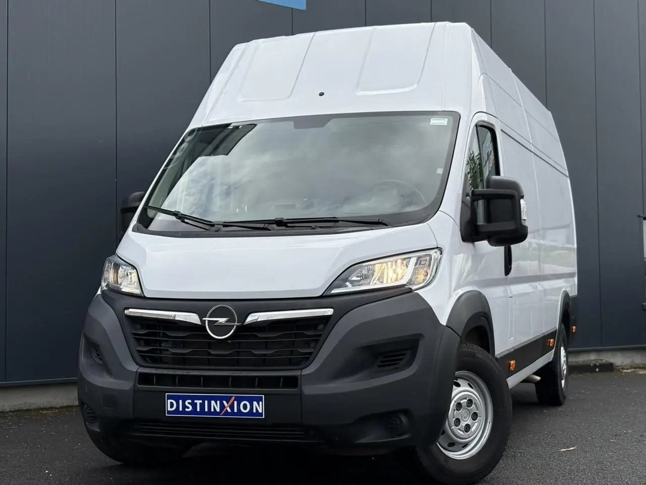 Opel Movano Fourgon blanc en 3/4 avant droit avec calandre noire et feux allumés sur fond urbain.
