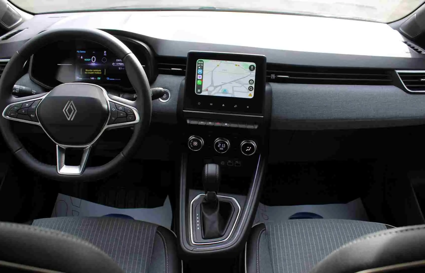 Vue intérieure avant de la Renault Clio 2025 hybride, tableau de bord gris noir avec écran tactile central et volant multifonction.