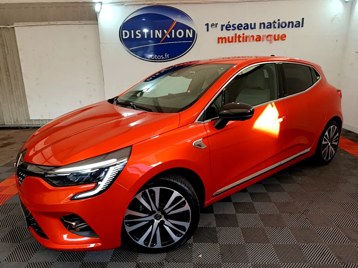 Vue 3/4 avant droite d'une Renault Clio Initiale Paris orange vif dans un showroom avec sol damier noir et rouge.