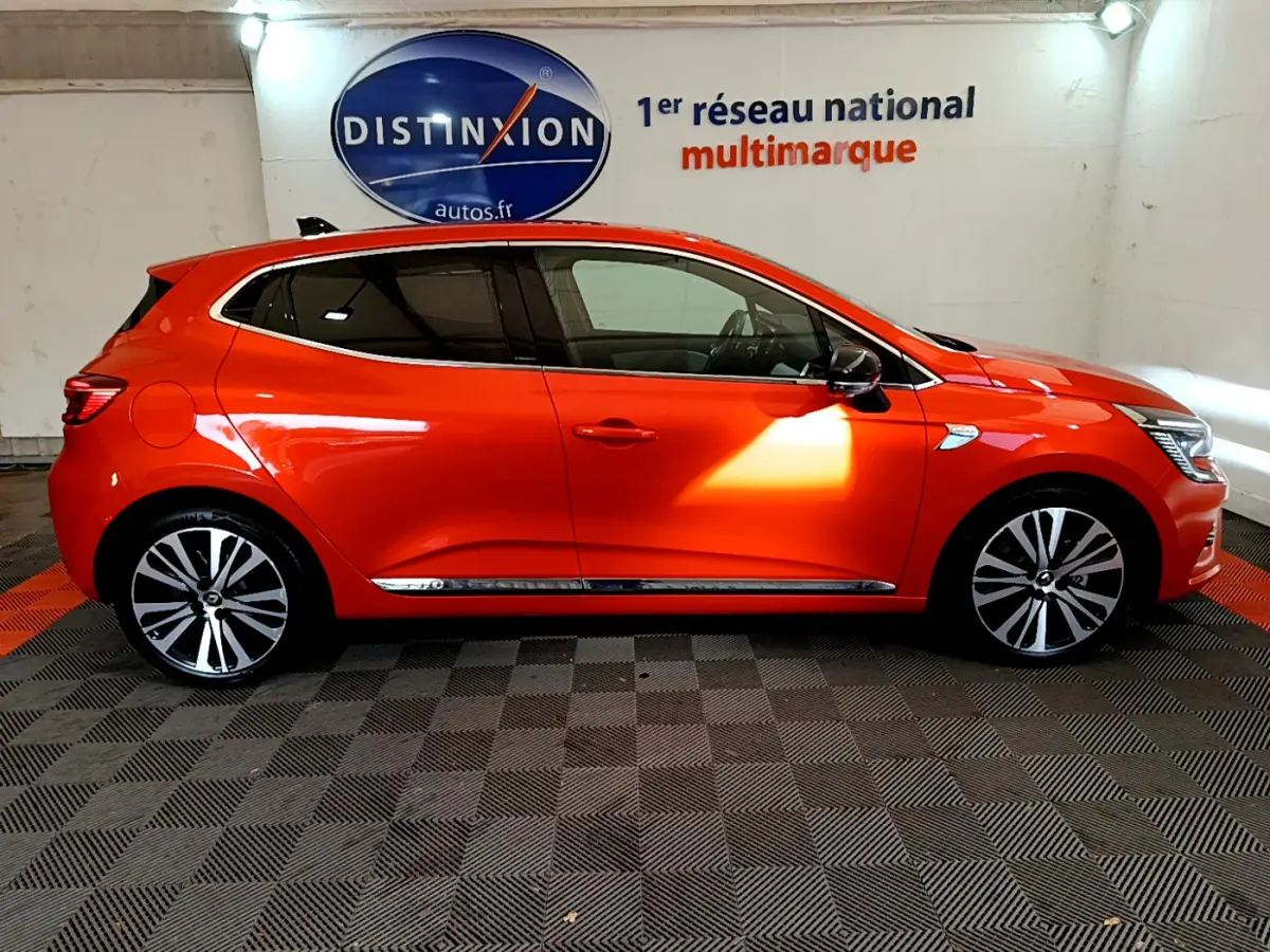 Profil côté droit d'une Renault Clio Initiale Paris orange vif, avec jantes alliage et vitres teintées en intérieur showroom.