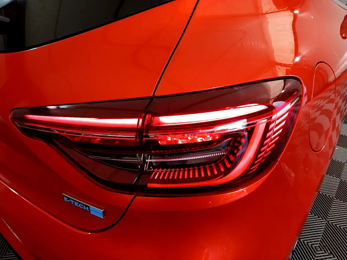 Gros plan sur le feu arrière droit allumé d'une Renault Clio Initiale Paris E-TECH orange 2021 avec logo hybride visible.