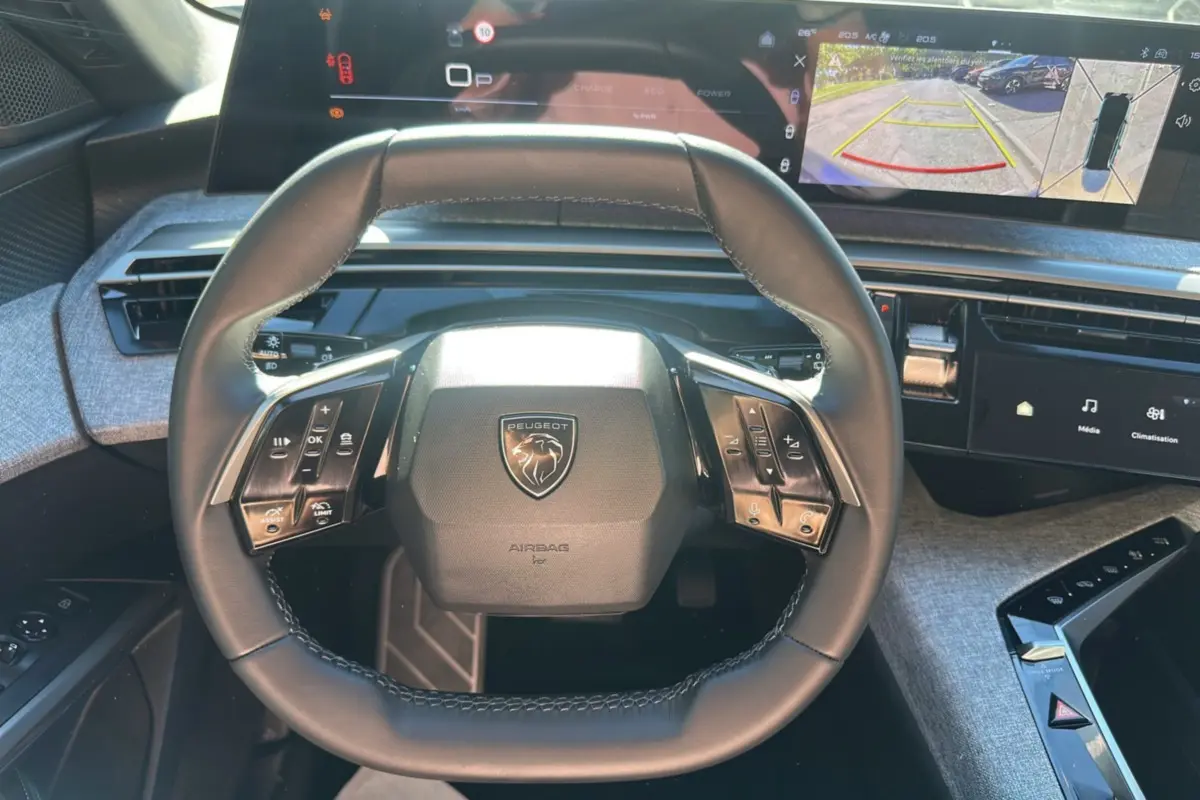 Vue rapprochée du volant cuir multifonctions Peugeot 5008 Hybrid 2025 avec tableau de bord numérique et écran caméra 360° affichés.