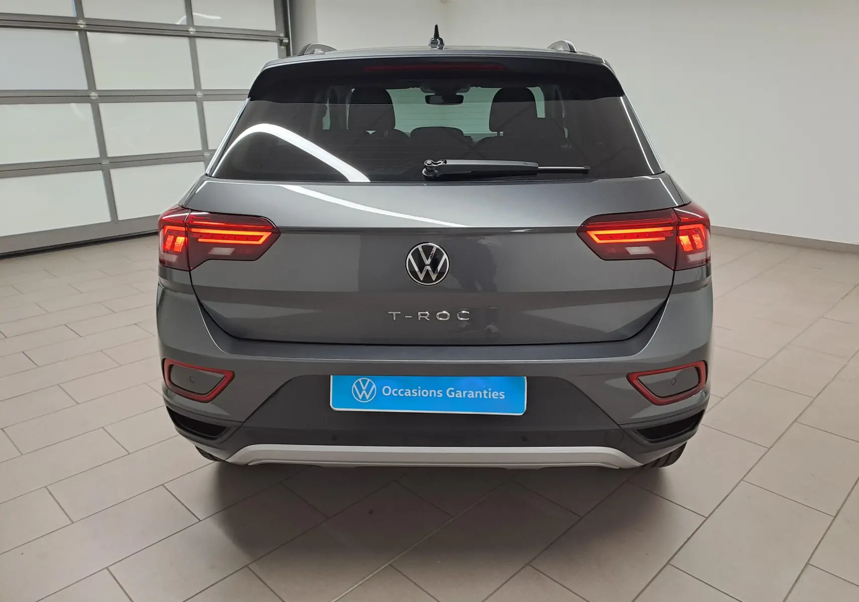 Vue arrière d'un Volkswagen T-Roc 2024 gris Indium avec feux allumés et plaque "Occasions Garantis" en intérieur showroom.