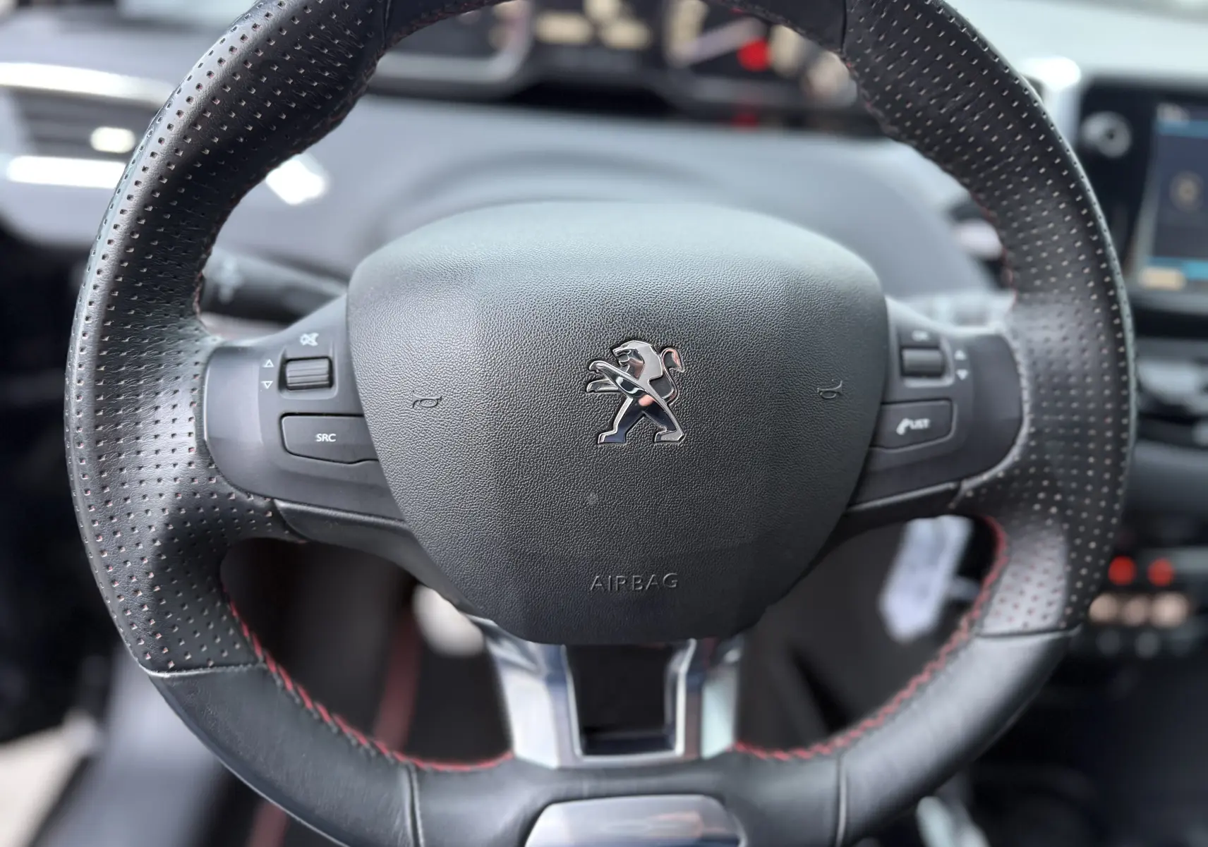 Gros plan sur le volant perforé avec surpiqûres rouges du Peugeot 2008 GT Line noir, tableau de bord flou en arrière-plan.