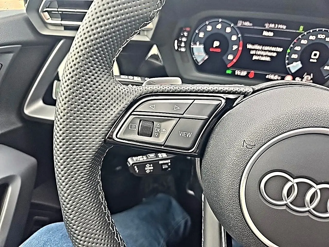 Gros plan sur le volant cuir perforé côté gauche de l'Audi A3 Sportback noir mythos avec tableau de bord digital.