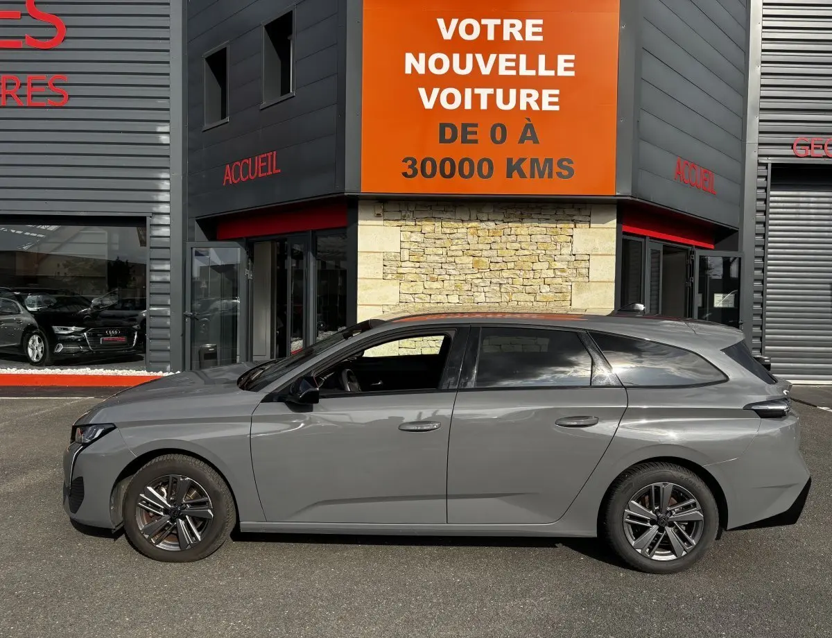 Profil côté gauche d'une Peugeot 308 PureTech 130 S&S Allure gris clair 2024 stationnée devant un bâtiment commercial.