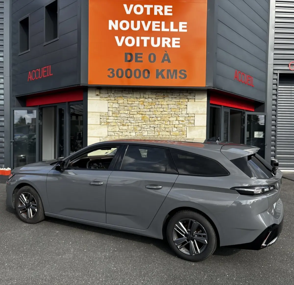 Peugeot 308 PureTech 130 gris clair vue de profil côté gauche, break garé devant un bâtiment avec panneau orange.
