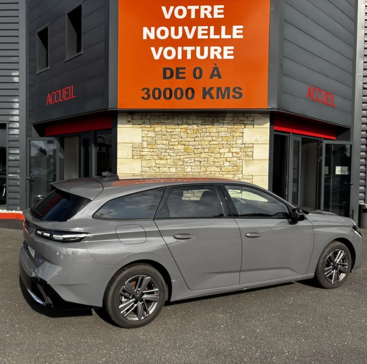 Peugeot 308 PureTech 130 S&S Allure gris clair vue de profil côté gauche devant un bâtiment avec panneau orange "Votre nouvelle voiture".