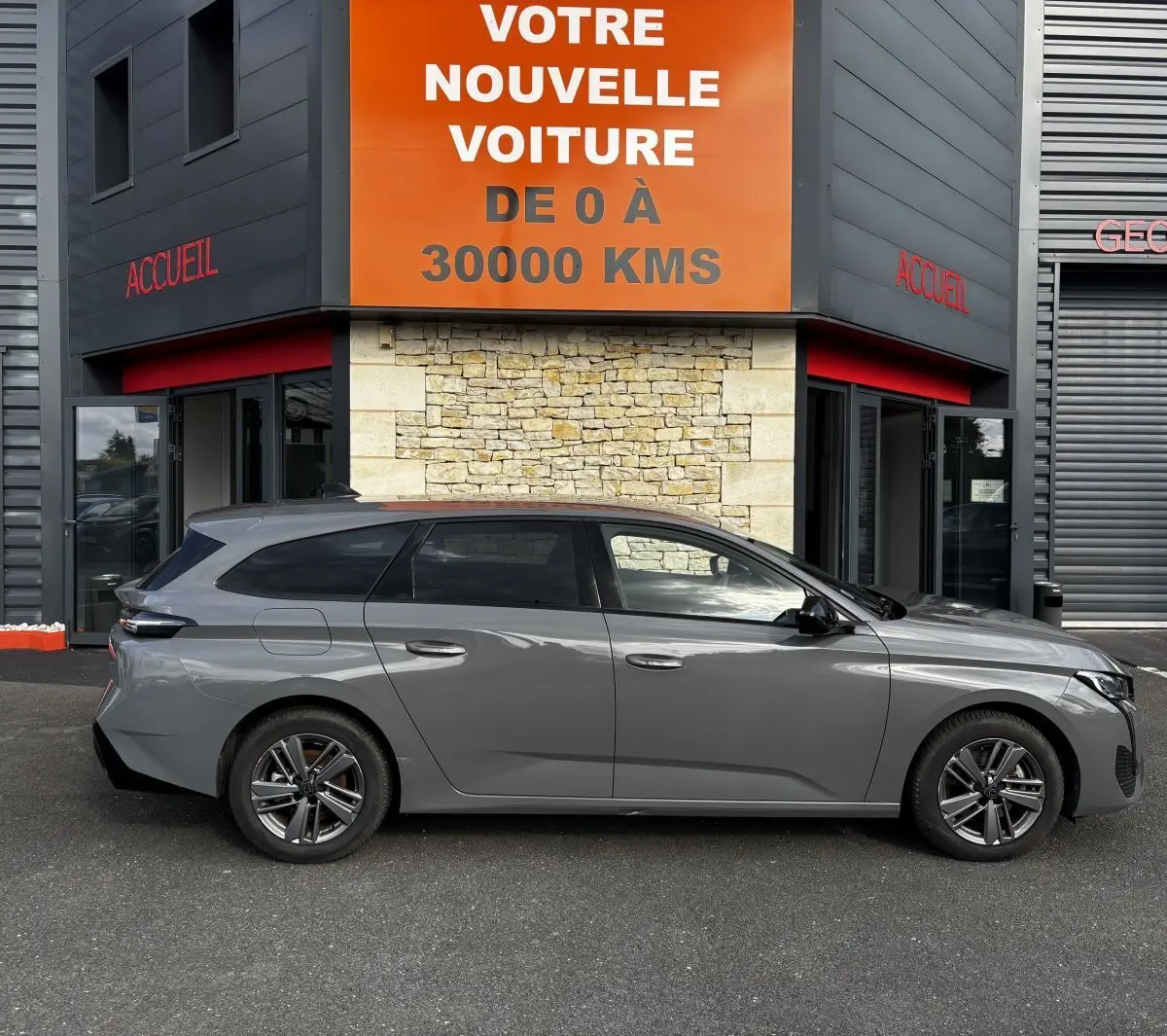 Peugeot 308 PureTech 130 S&S Allure gris clair vue de profil côté gauche devant un bâtiment avec panneau orange.