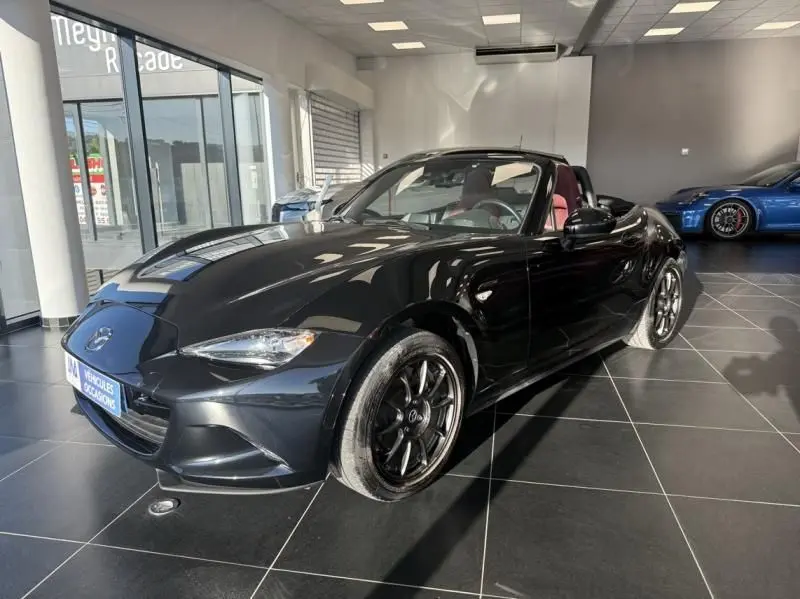 Mazda MX-5 noire vue en 3/4 avant droit dans un showroom, capote ouverte et intérieur rouge visible.