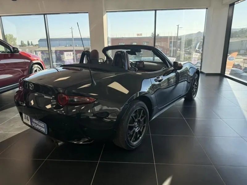 Mazda MX-5 noir 2020 en 3/4 arrière droit, capote ouverte, jantes noires et intérieur visible en showroom.