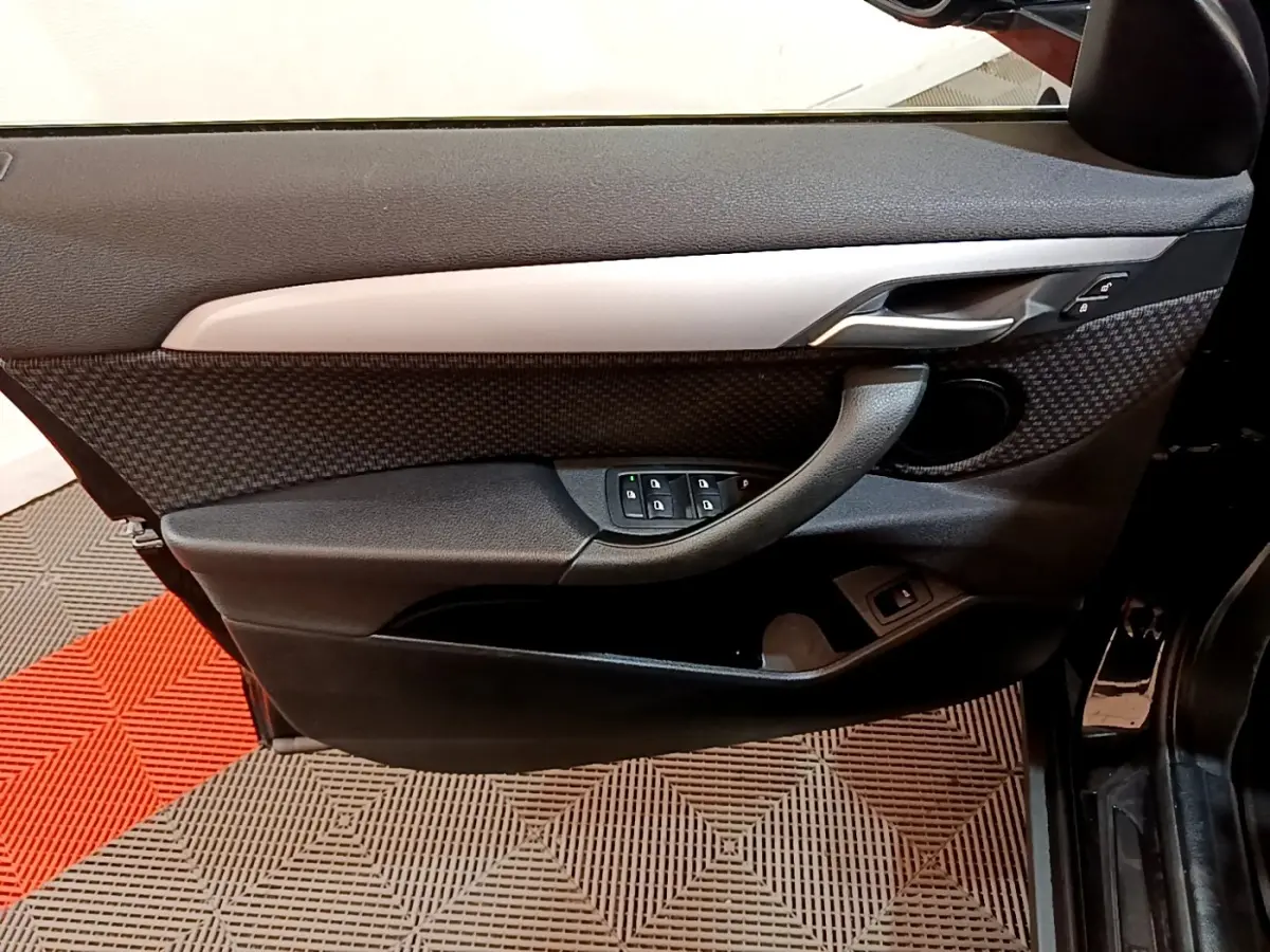Vue intérieure côté gauche de la porte avant noire de la BMW X2 sDrive18i Lounge avec commandes de vitres et poignée intégrée.