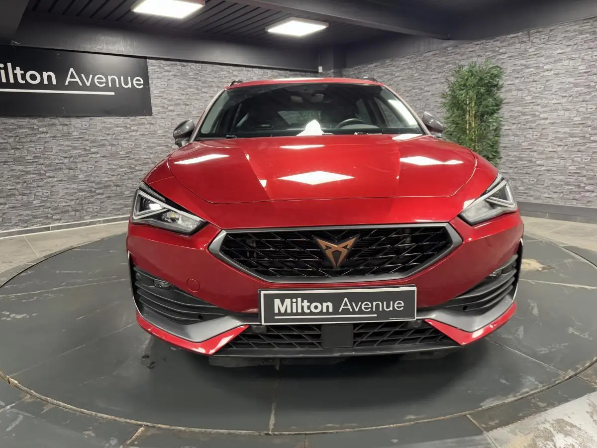 Vue frontale d'une CUPRA Leon Sportstourer rouge Désir avec calandre noire et logo cuivré dans un showroom.