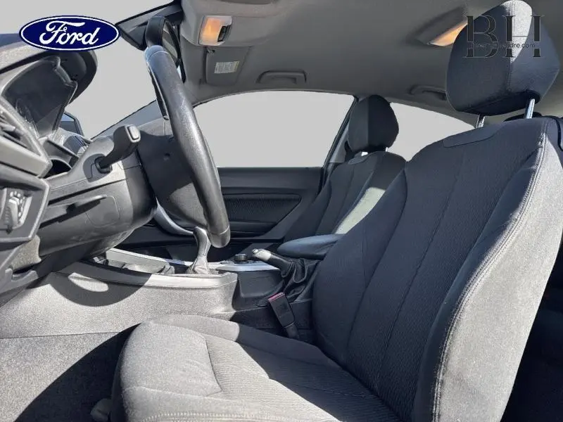 Vue intérieure côté conducteur d'une BMW Série 1 114i 2013 avec sièges tissu noirs et volant cuir.