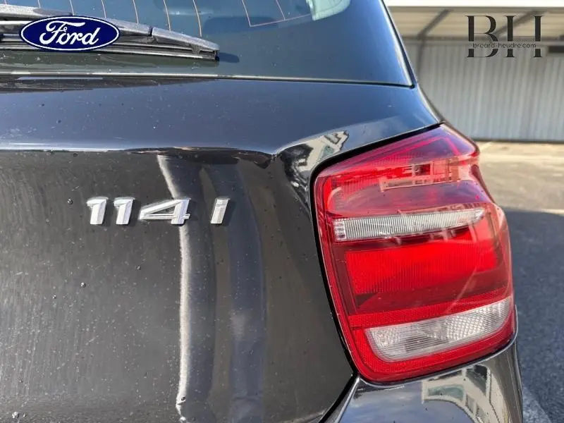 Gros plan sur le feu arrière droit et le badge 114i d'une BMW Série 1 noire, vue arrière côté droit.