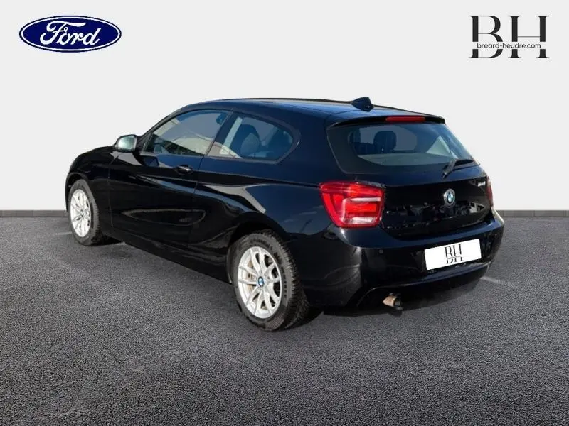 Vue 3/4 arrière droite d'une BMW Série 1 114i noire, avec jantes alu 16 pouces et feux arrière rouges.