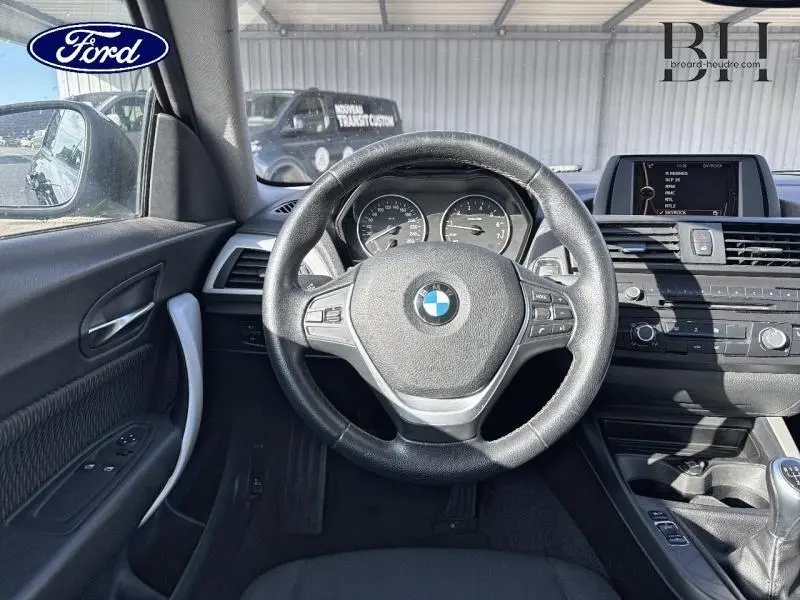Vue intérieure centrée sur le volant cuir de la BMW Série 1 114i noire avec tableau de bord et écran multifonction.