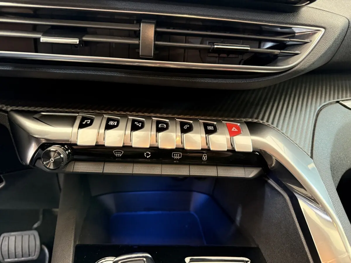 Gros plan sur la console centrale du Peugeot 3008 bleu, mettant en valeur les boutons de commande en métal et le revêtement carbone.