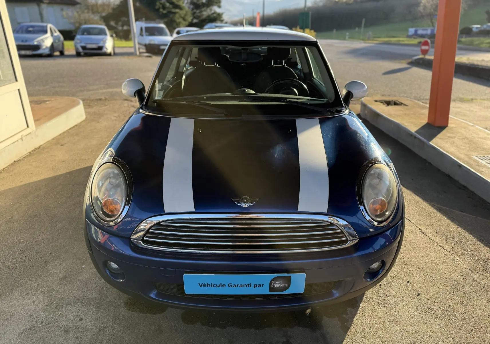 Vue avant d'une MINI Cooper bleu avec deux bandes blanches sur le capot et calandre chromée sous un soleil bas