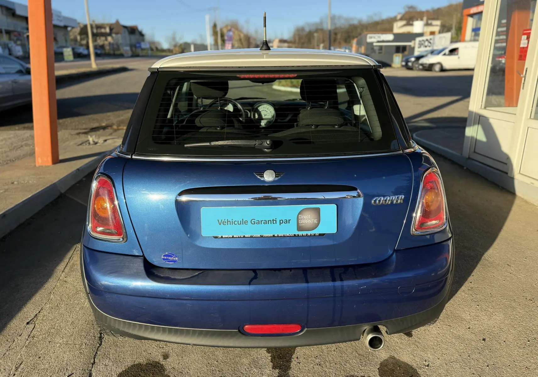 Vue arrière d'une MINI Cooper bleu avec toit blanc, feux arrière allumés et plaque d'immatriculation bleue.