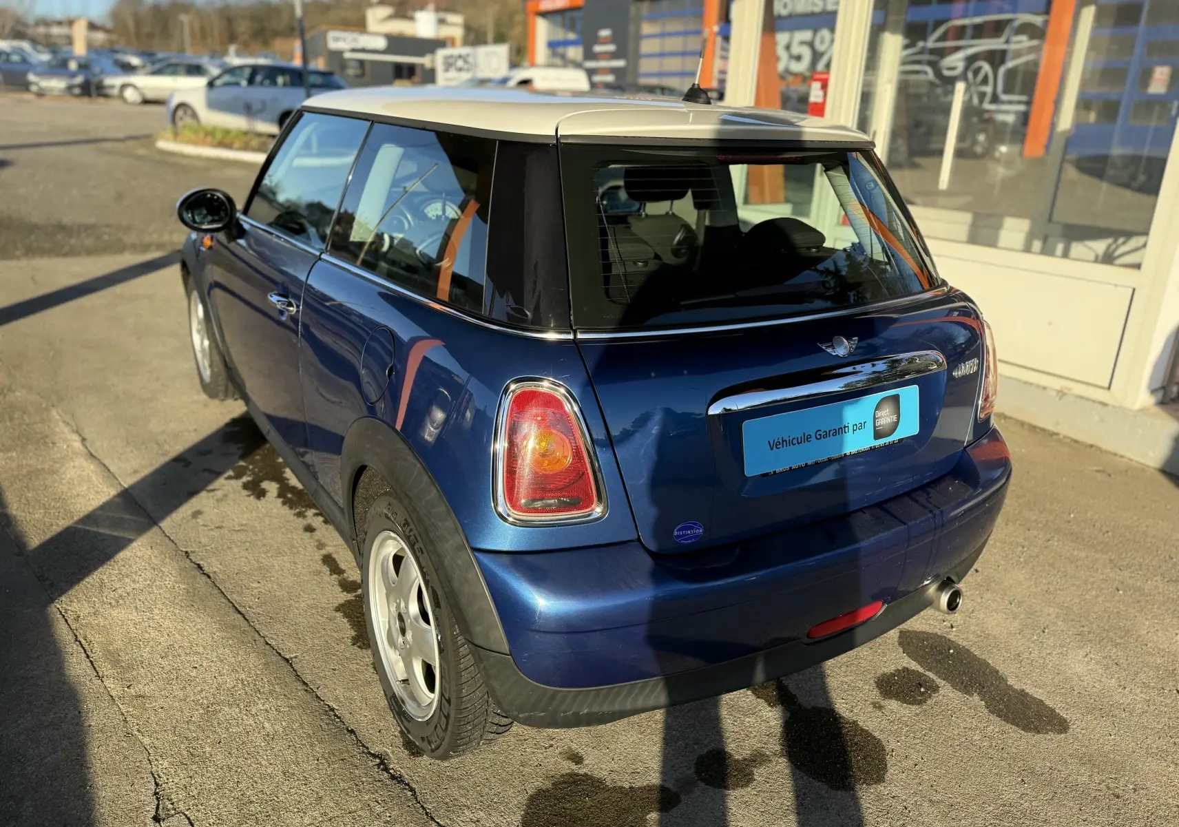 Vue 3/4 arrière droite d'une MINI Cooper bleu avec toit blanc garée devant un bâtiment commercial.