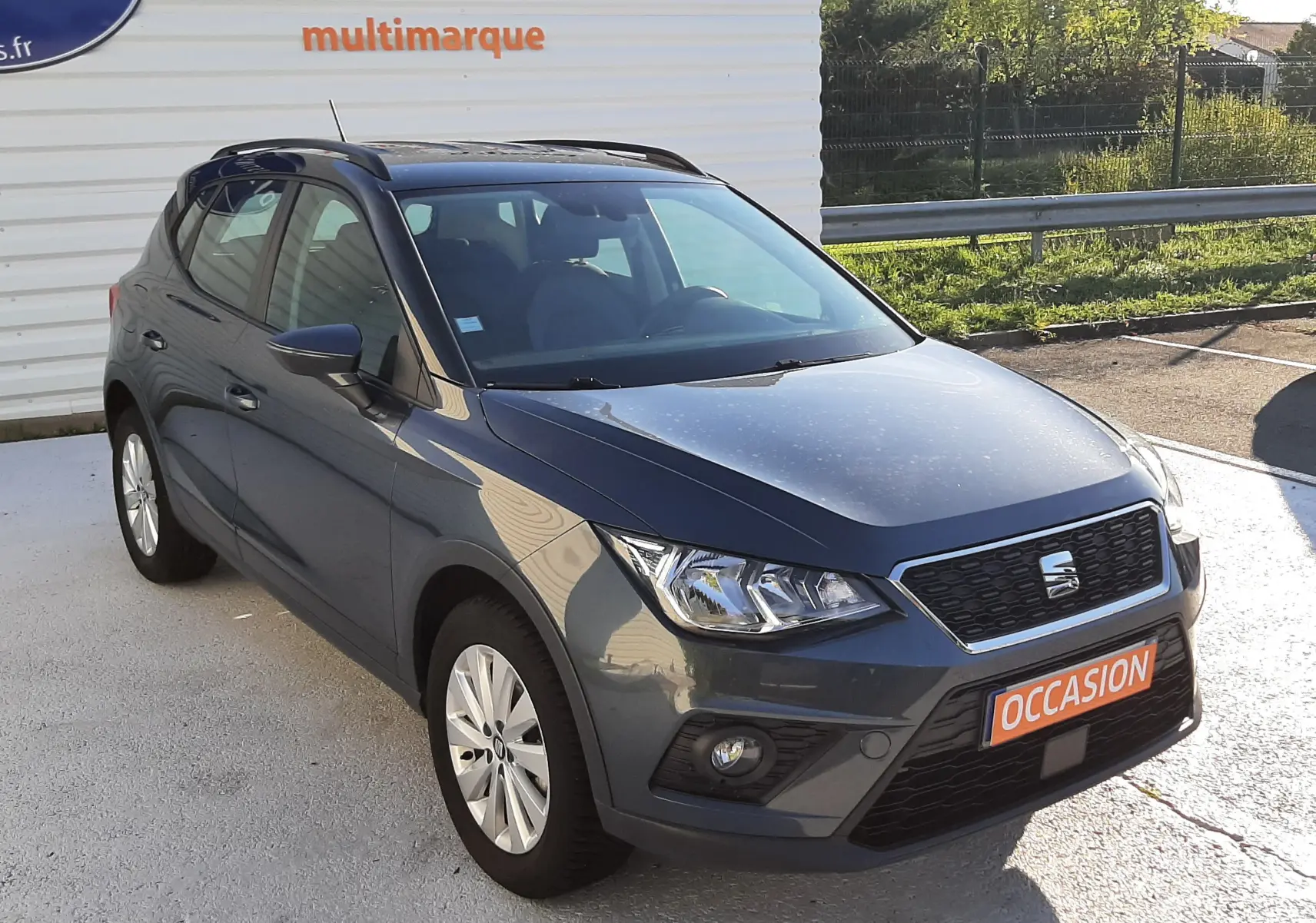 SEAT Arona gris Magnetic avec toit noir, vue 3/4 avant droit, sur parking devant panneau publicitaire.