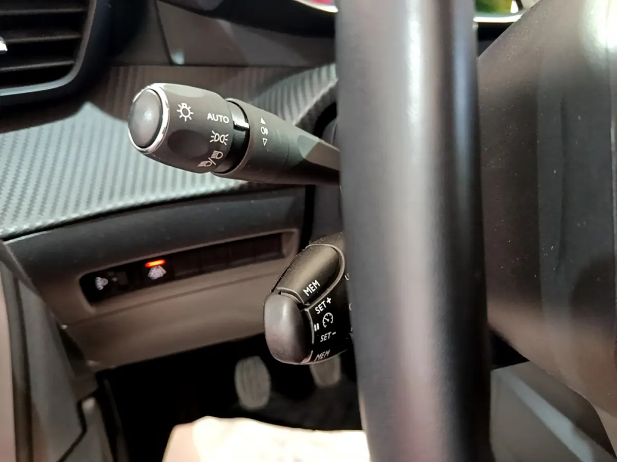 Gros plan sur les commandes de phares et régulateur de vitesse derrière le volant cuir d'une Peugeot 208 gris clair.