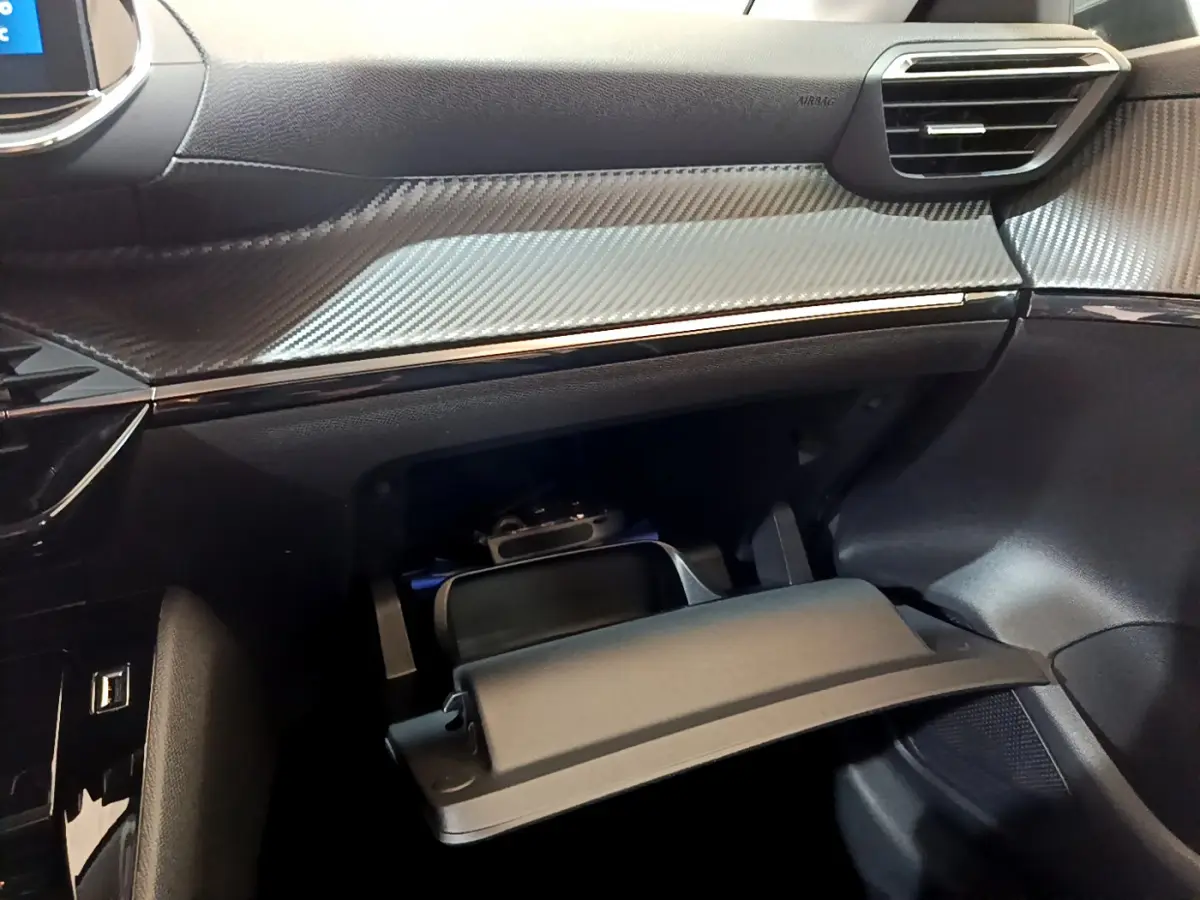 Vue rapprochée de la boîte à gants ouverte et tableau de bord noir avec insert effet carbone dans l'habitacle d'une Peugeot 208 2020.