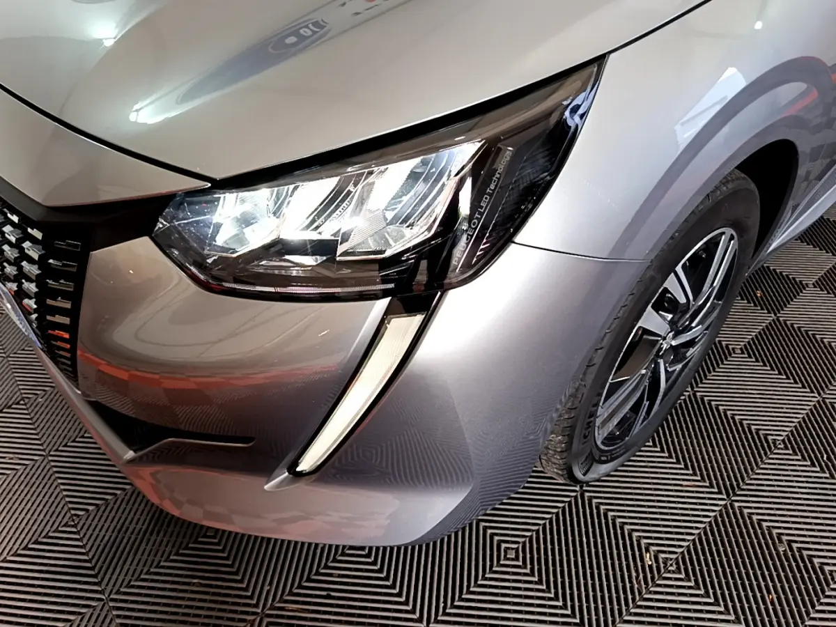Gros plan sur le phare avant droit et la jante d'une Peugeot 208 gris clair, version PureTech 100 S&S Allure Business.