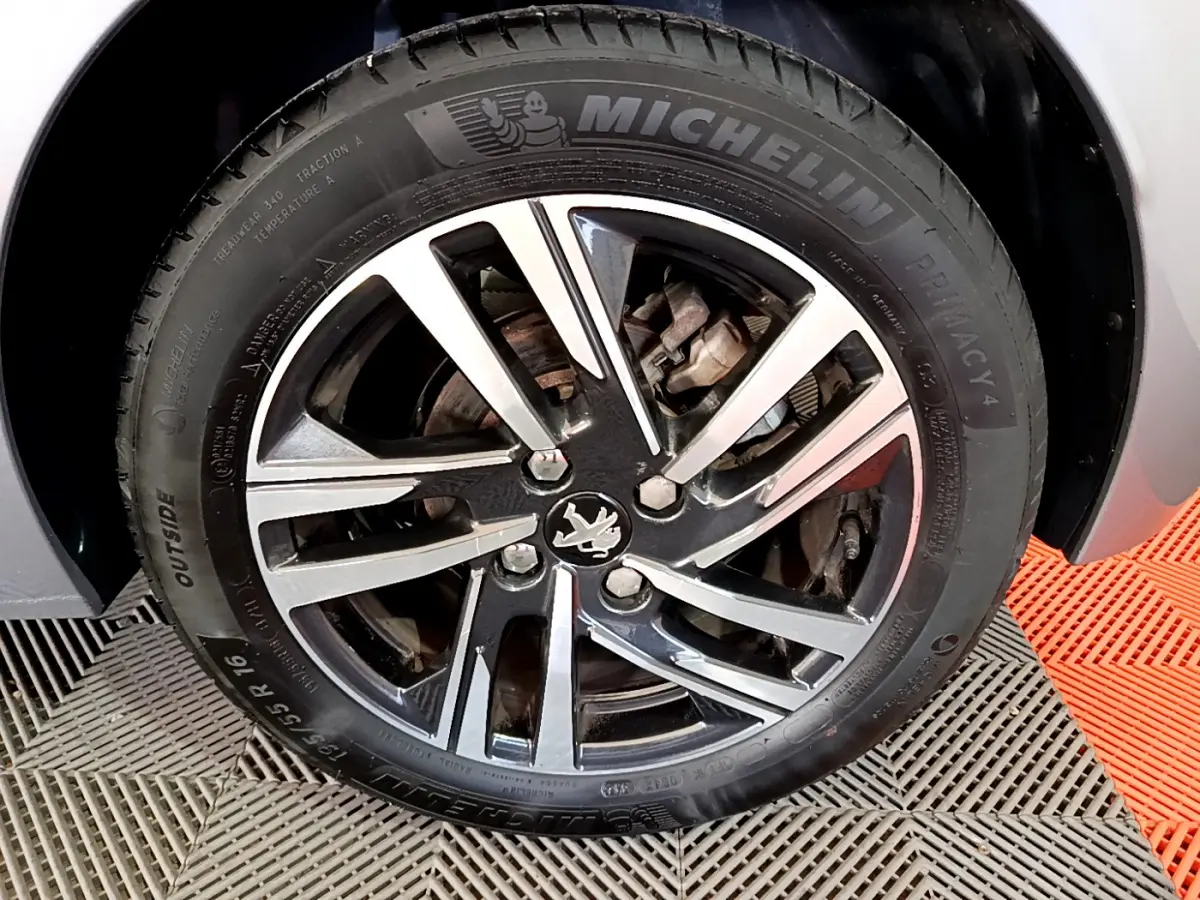 Gros plan sur la roue avant droite d'une Peugeot 208 gris clair, jante alliage noire et argent avec logo lion central.