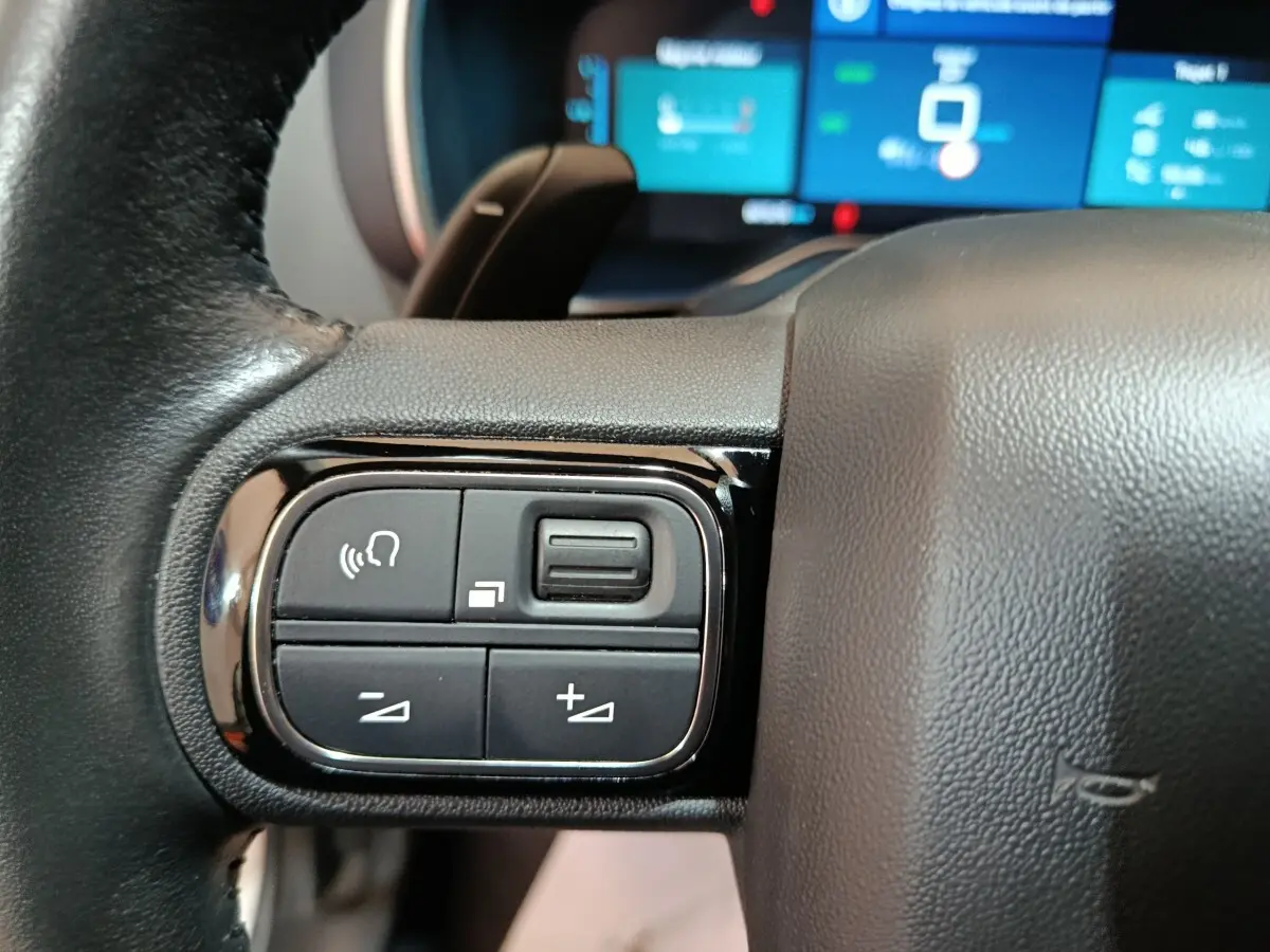 Gros plan sur les commandes multifonctions à gauche du volant du Citroën C5 Aircross Hybrid blanc, avec écran tactile en arrière-plan flou.