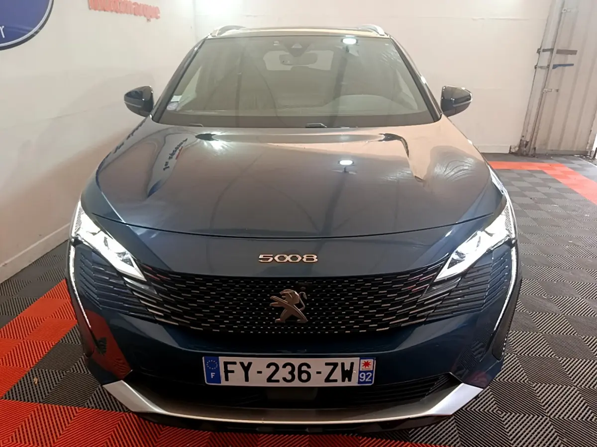 Vue avant d'un Peugeot 5008 bleu 2021 avec calandre noire et phares LED allumés dans un garage.