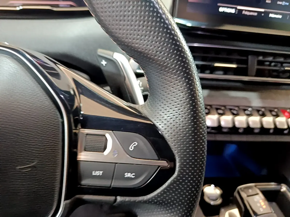 Gros plan sur le volant cuir perforé côté gauche du Peugeot 5008 bleu, avec commandes audio et tableau de bord en arrière-plan.