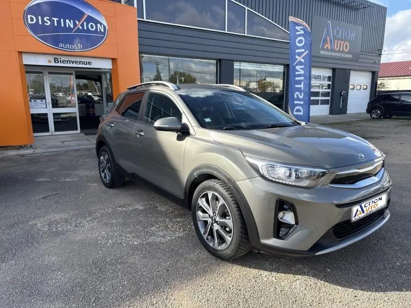 KIA Stonic brun Sienne vue 3/4 avant droit, avec barres de toit et jantes alliage, stationné devant un garage.