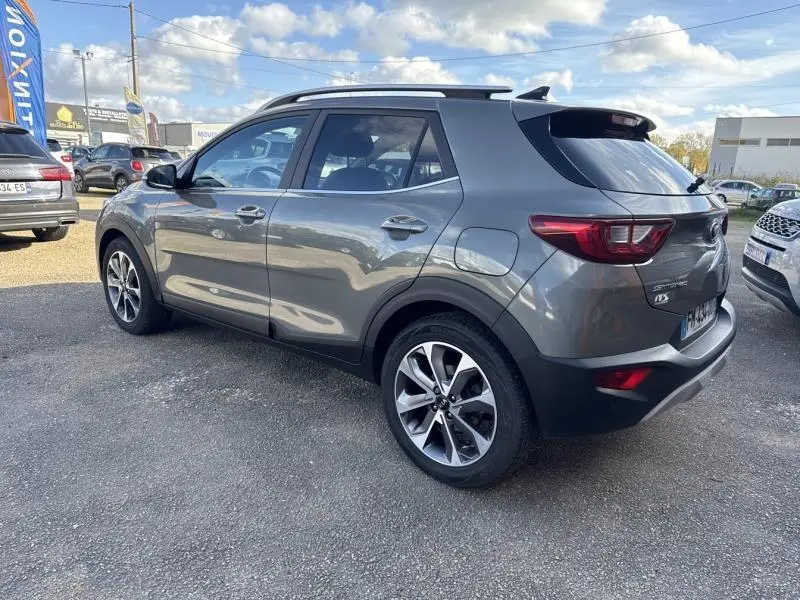 Vue 3/4 arrière droite d'un KIA Stonic gris métallisé avec jantes alliage et barres de toit sur parking extérieur.