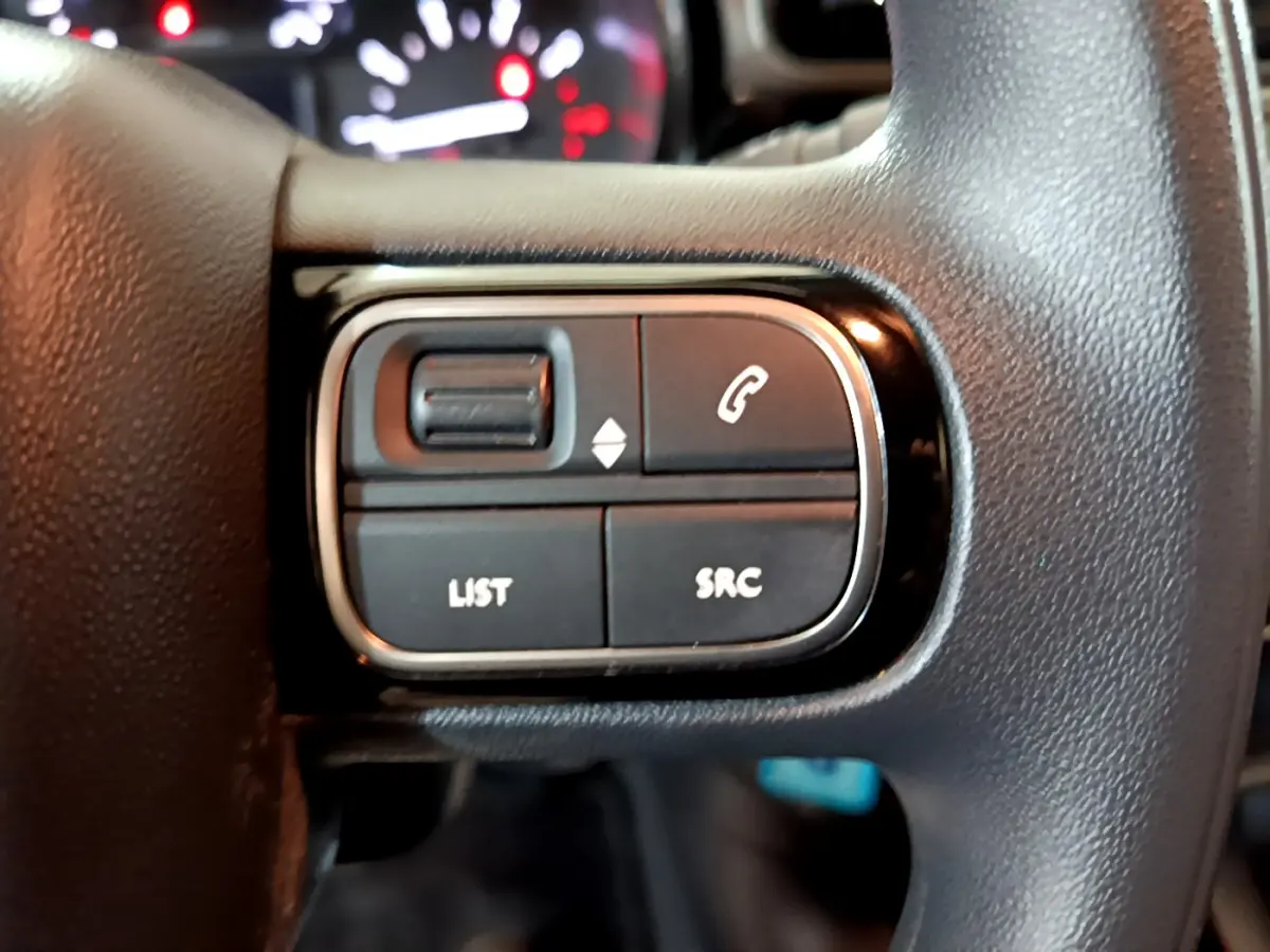 Gros plan sur les commandes multimédia noires du volant d'une Citroën C3 beige 2022, avec tableau de bord flou en arrière-plan.