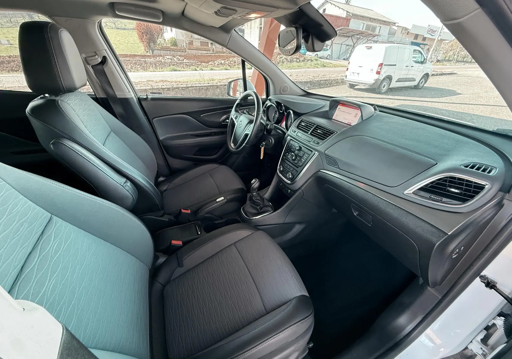 Intérieur de l'Opel Mokka X 2016 vu côté conducteur, sièges tissu gris et tableau de bord noir avec écran central.