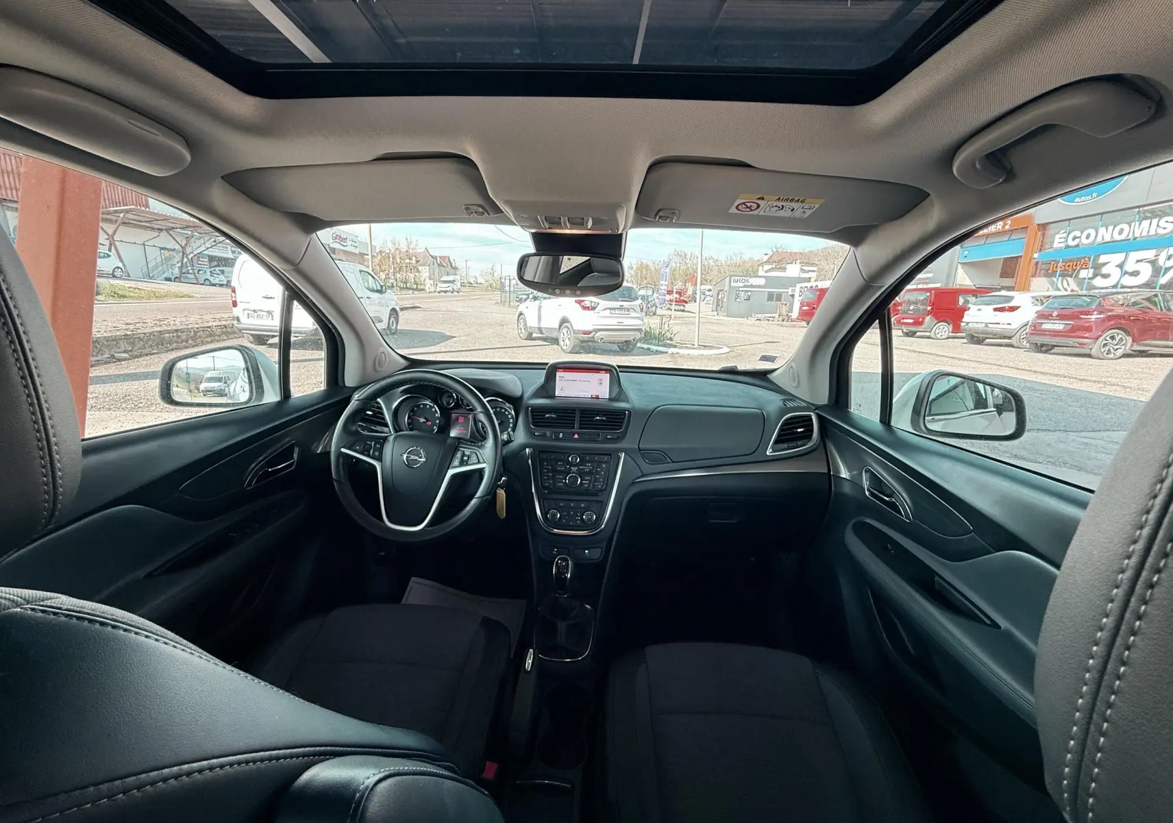 Vue intérieure avant de l’Opel Mokka X blanc, montrant le tableau de bord, le volant et le toit ouvrant panoramique.