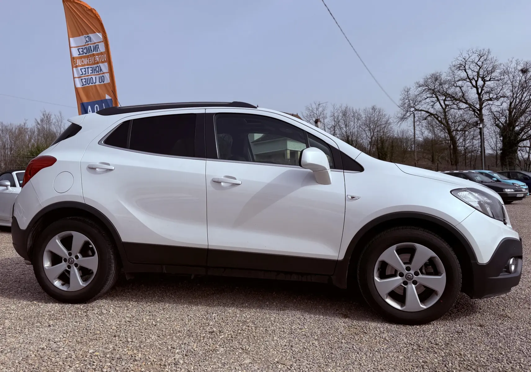 Vue de profil côté gauche d'un Opel Mokka X blanc avec jantes alliage et toit noir sur un parking extérieur.