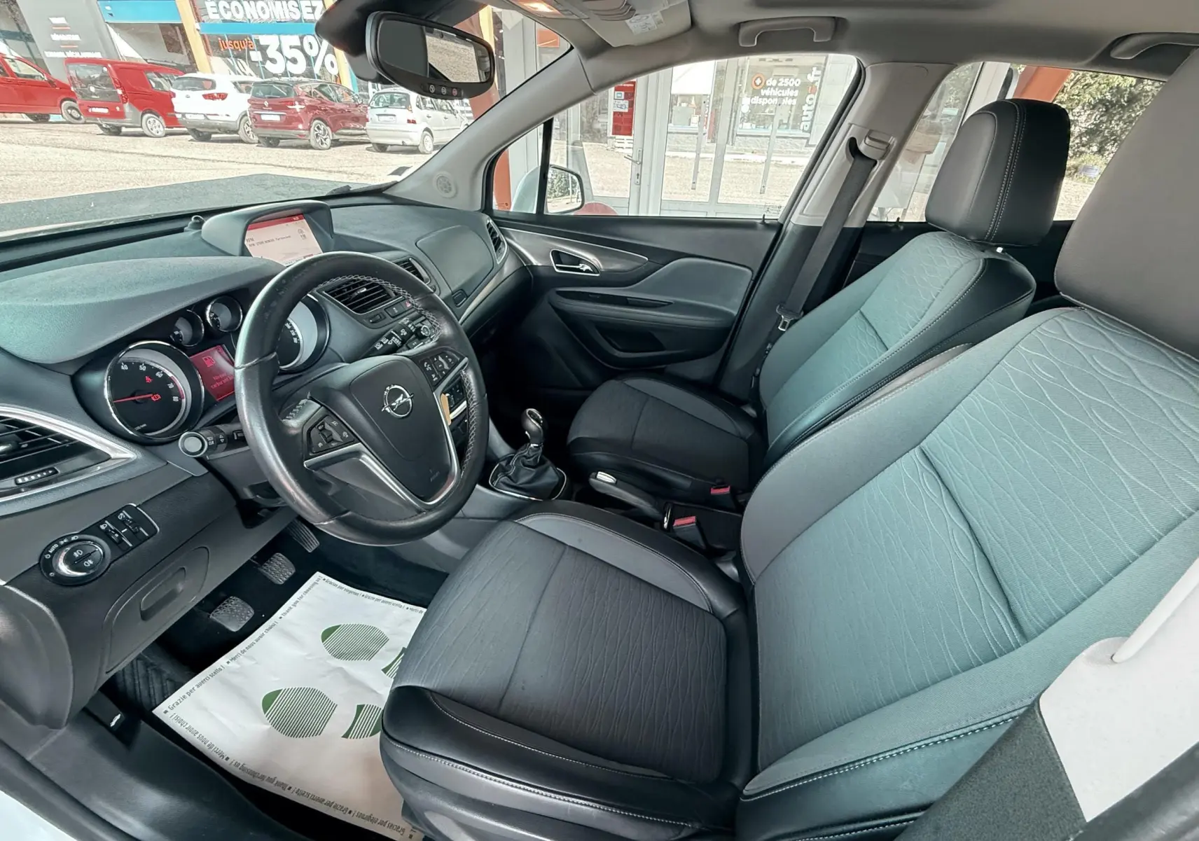 Vue intérieure côté conducteur de l'Opel Mokka X 2016, sièges tissu gris et cuir noir, volant multifonction et tableau de bord visible.