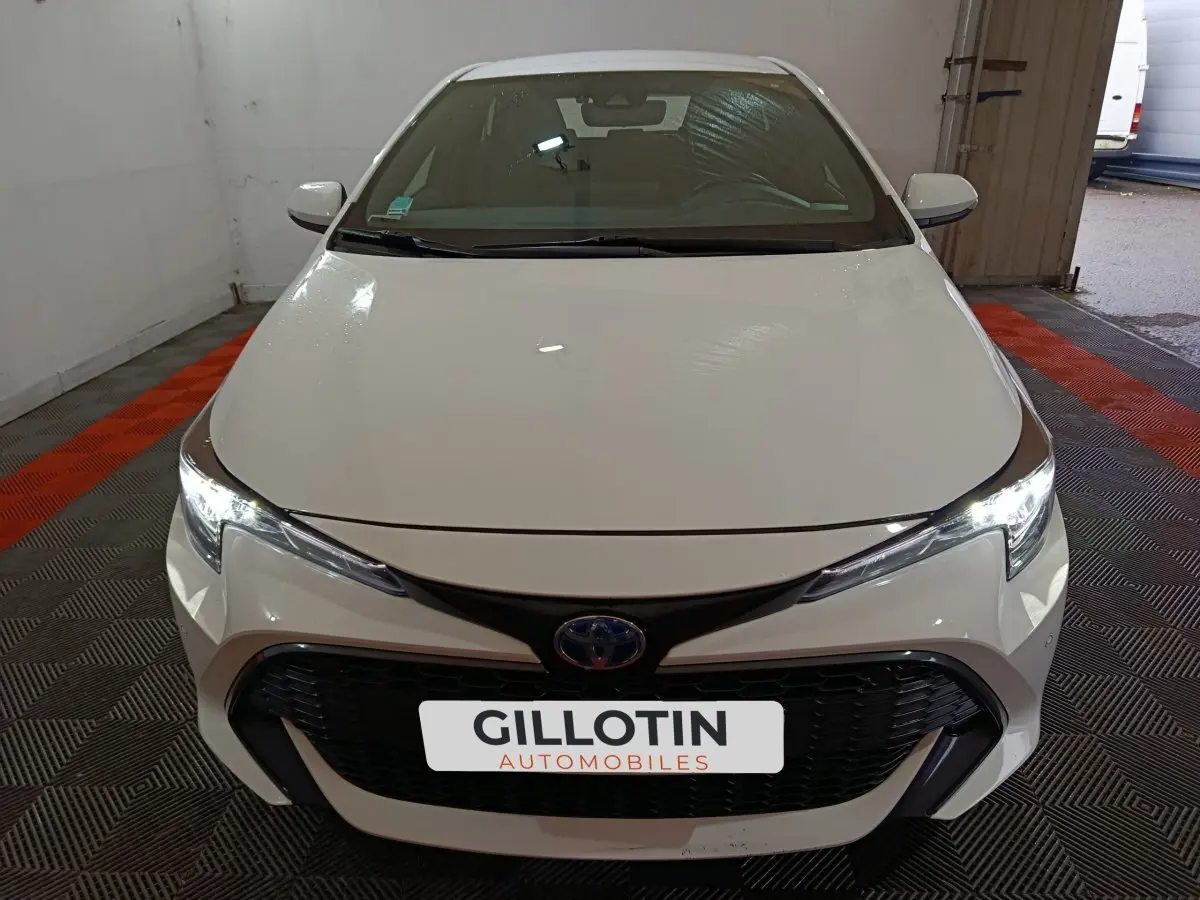 Vue avant d'une Toyota Corolla hybride blanche 2022 avec calandre noire et phares LED allumés dans un garage.