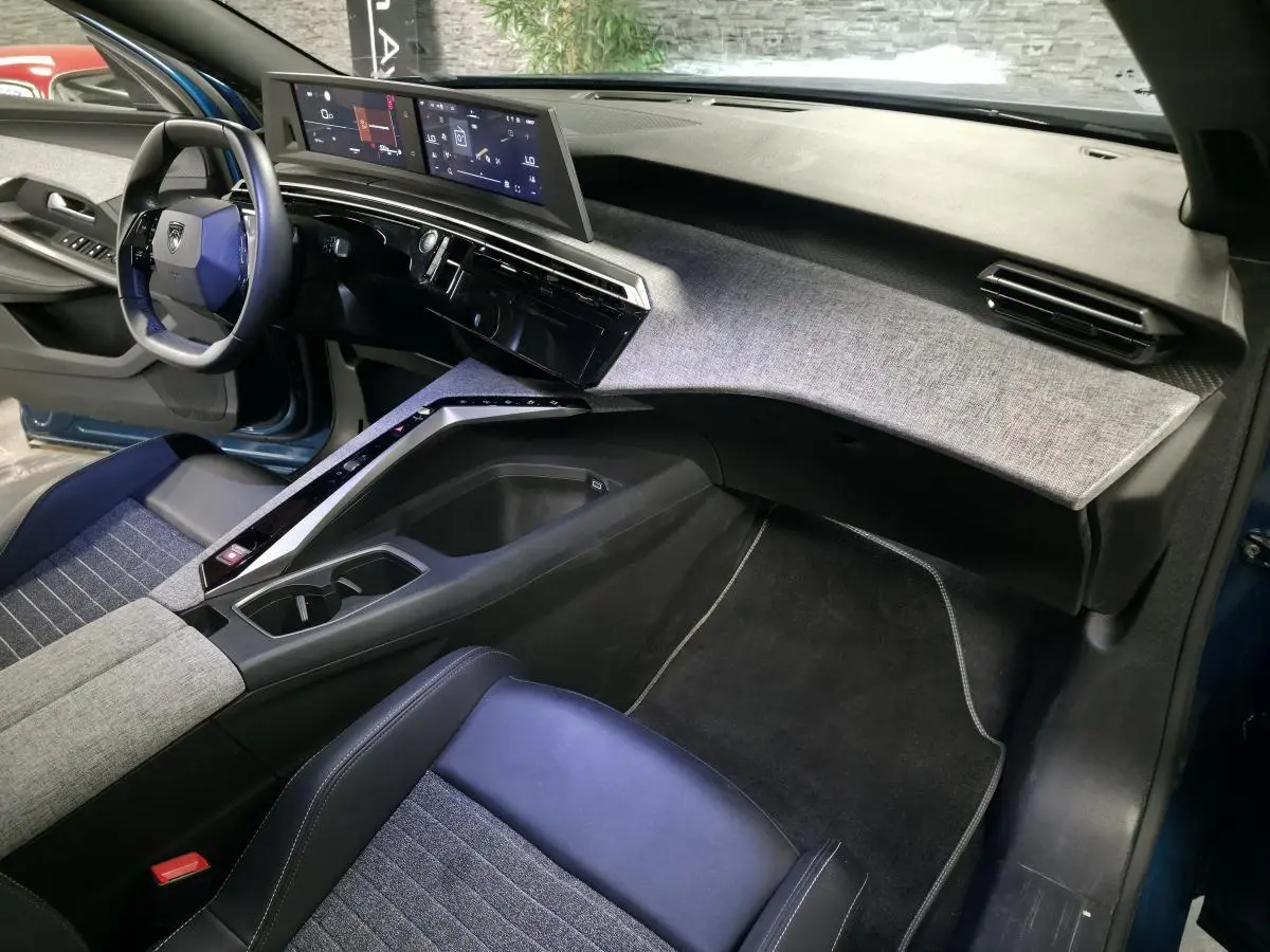 Vue intérieure côté passager du Peugeot 5008 bleu 2025, avec tableau de bord et console centrale au décor tissu gris et noir laqué.
