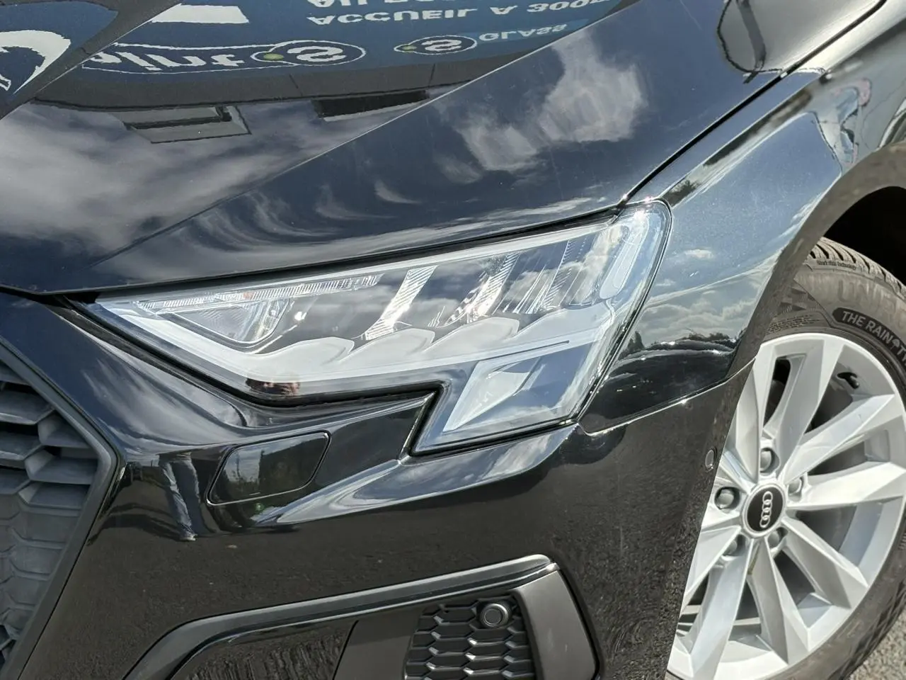 Gros plan sur le phare avant droit et la roue alliage 16 pouces d'une Audi A3 Sportback 30 TFSI noire Mythe 2023.