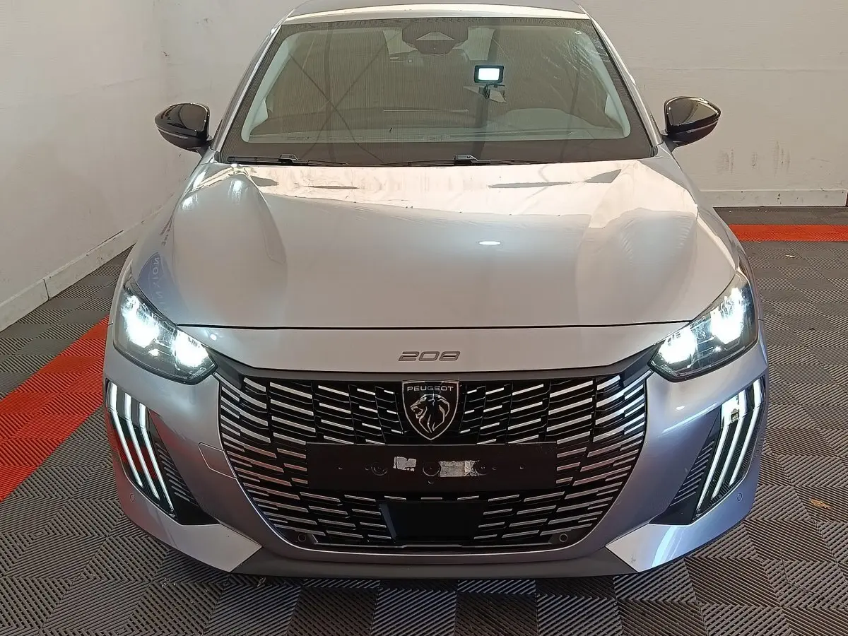 Vue frontale d'une Peugeot 208 gris clair 2025 avec phares LED allumés et calandre imposante au lion Peugeot.