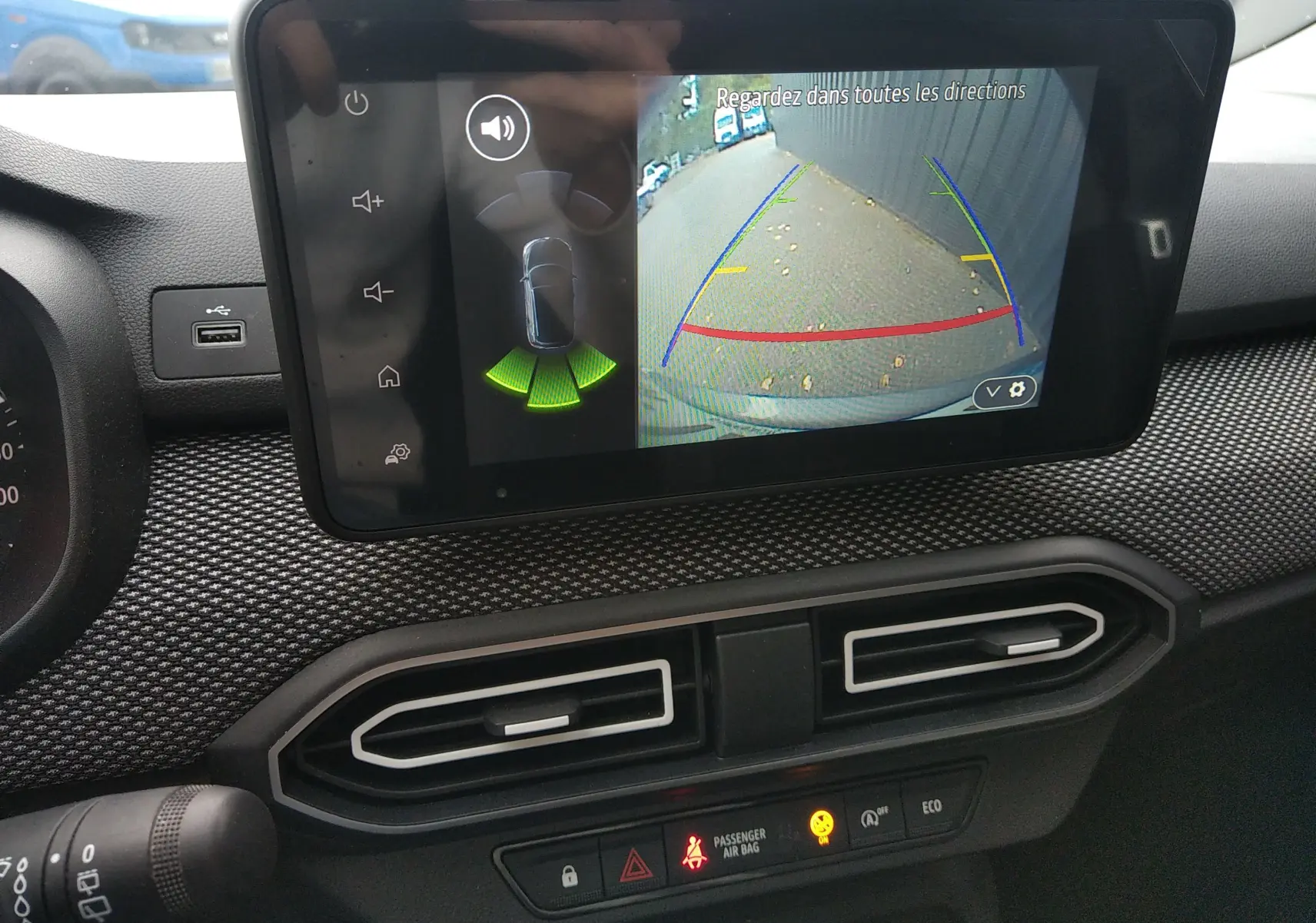 Écran central de la Dacia Sandero 2024 gris montrant la caméra de recul avec guidage de trajectoire et capteurs d’aide au stationnement.