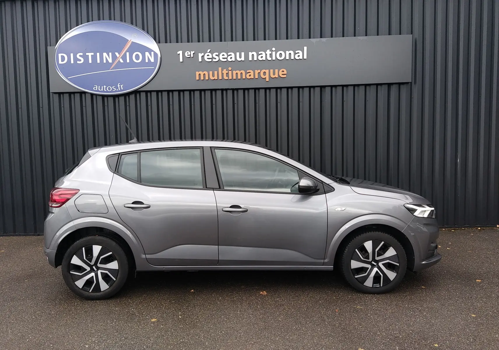 Vue latérale droite d'une Dacia Sandero gris métallisé 2024 garée devant un mur noir avec un panneau Distinxion.