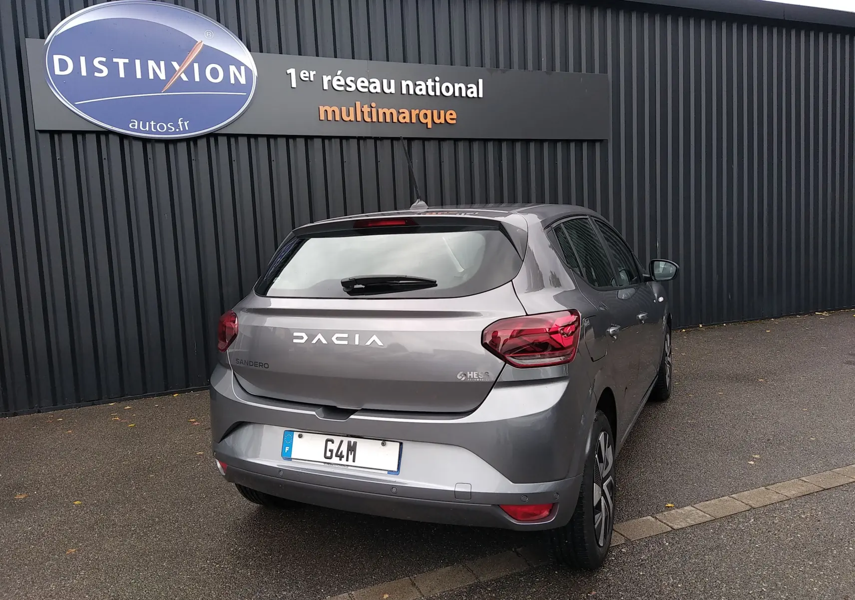 Vue 3/4 arrière droite d'une Dacia Sandero gris 2024 garée devant un mur noir avec enseigne Distinxion.
