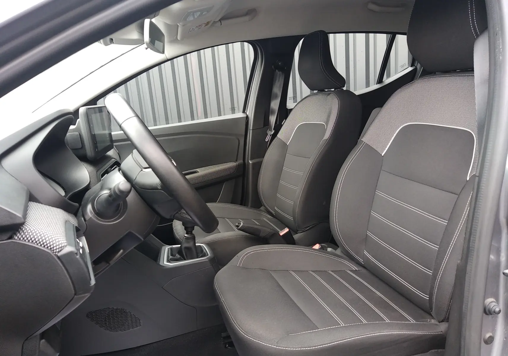 Vue intérieure côté conducteur de la Dacia Sandero 2024 gris, montrant les sièges tissu et la boîte manuelle.