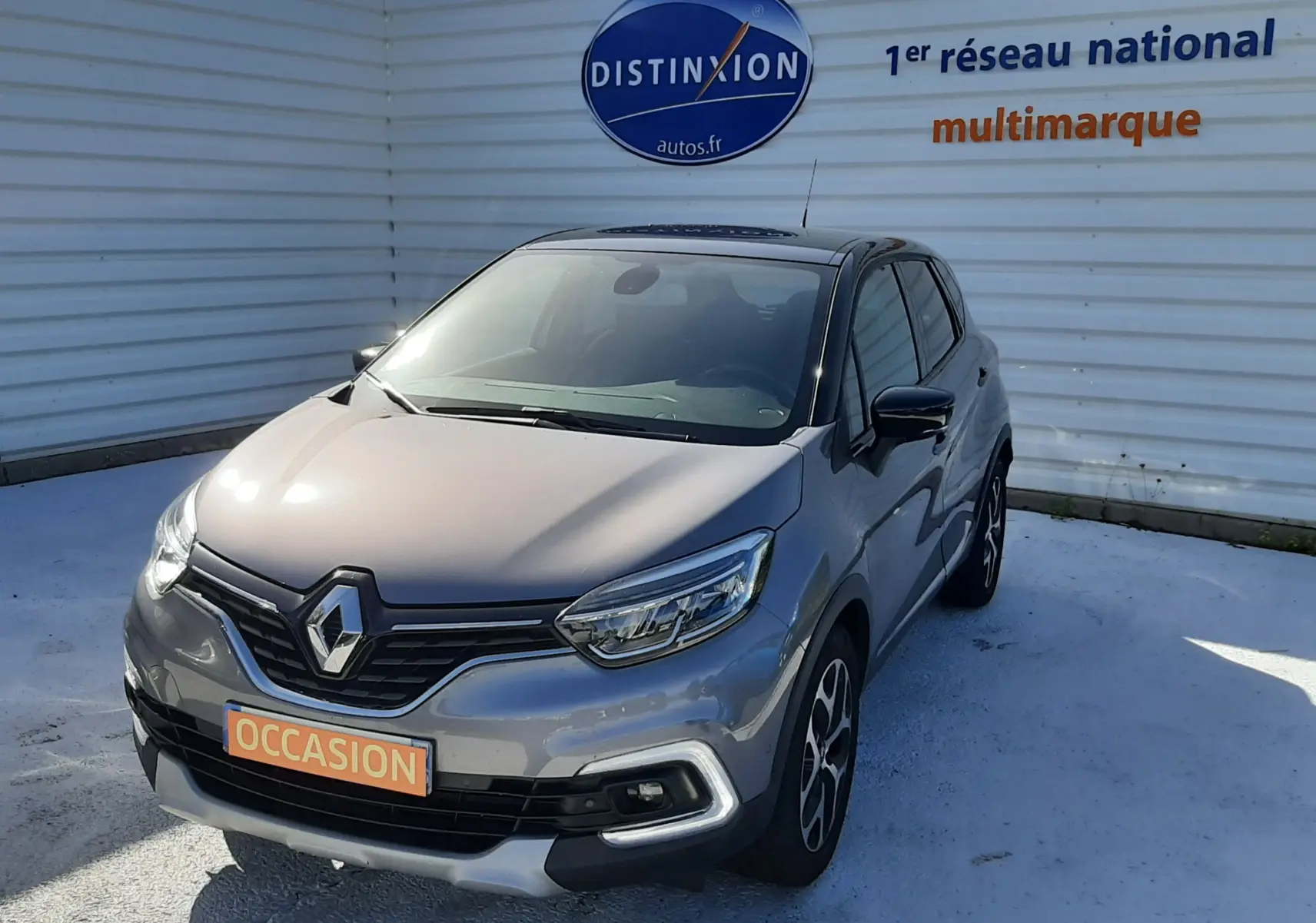 Renault Captur gris cassiope avec toit noir, vue 3/4 avant droit, sur parking devant un mur blanc marqué Distinxion.