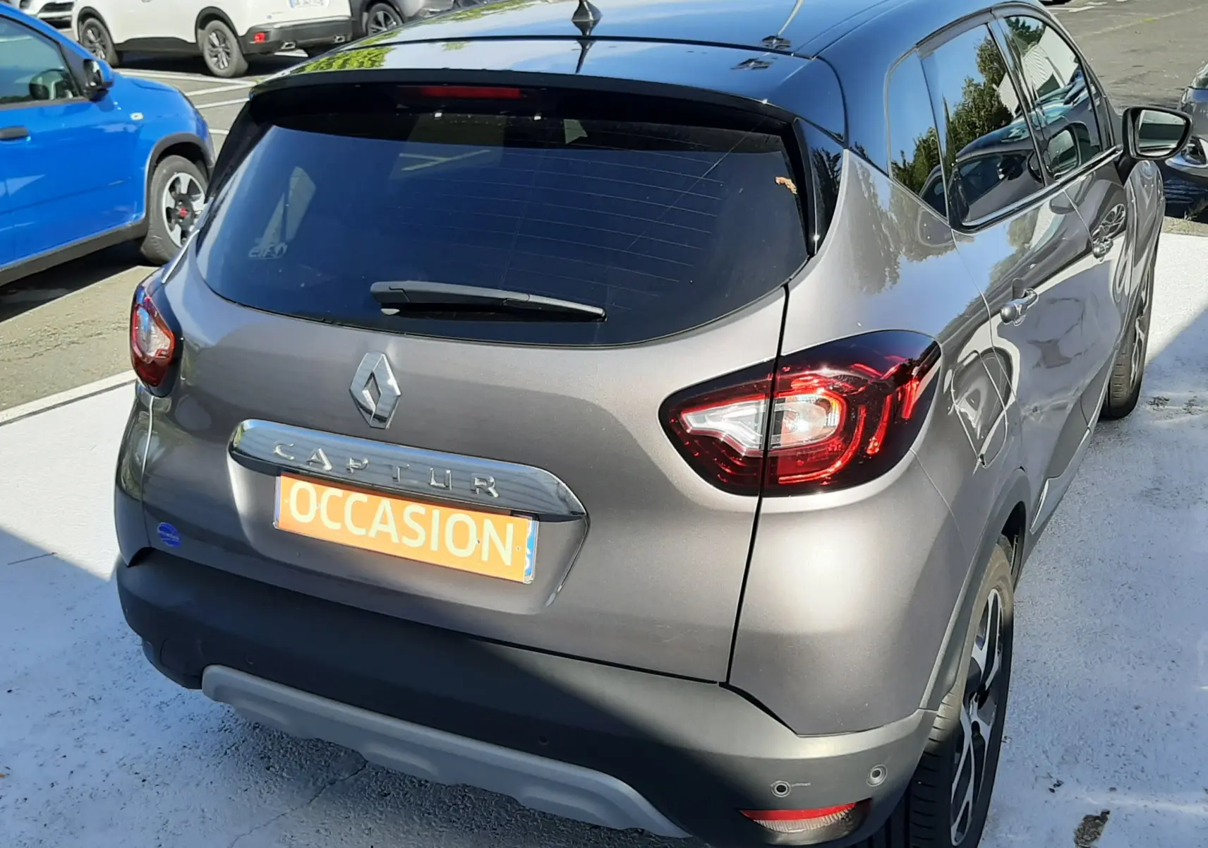 Vue 3/4 arrière droite d'un Renault Captur gris Cassiope avec toit noir, en vente sur parking automobile.